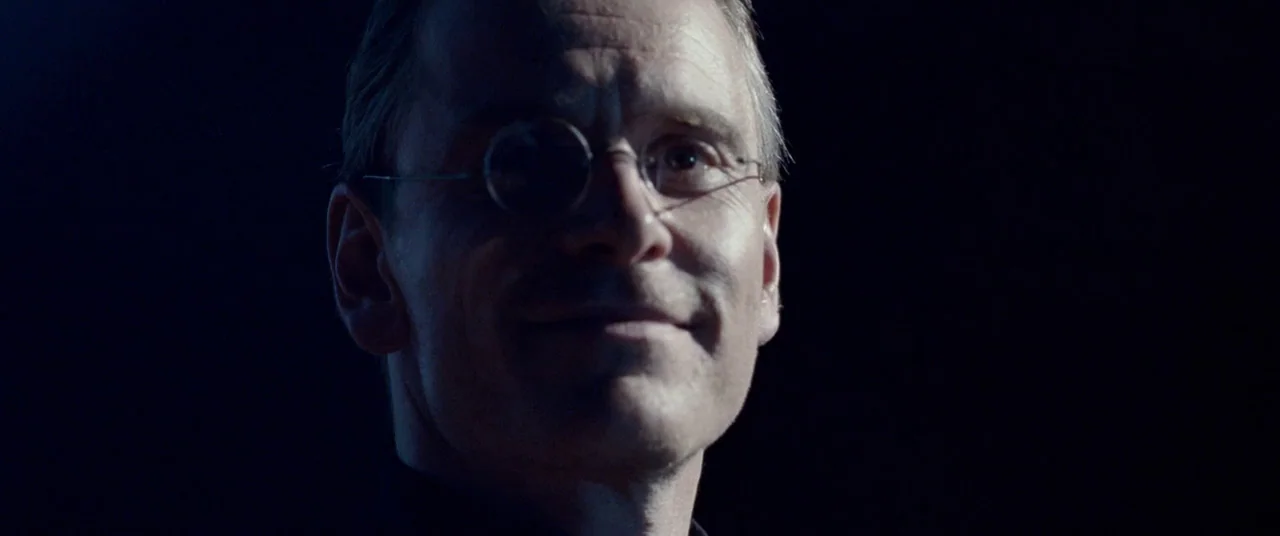 Steve Jobs (2015) movie still: closeup — A man stands in an underexposed environment with no background details visible. He looks directly toward…