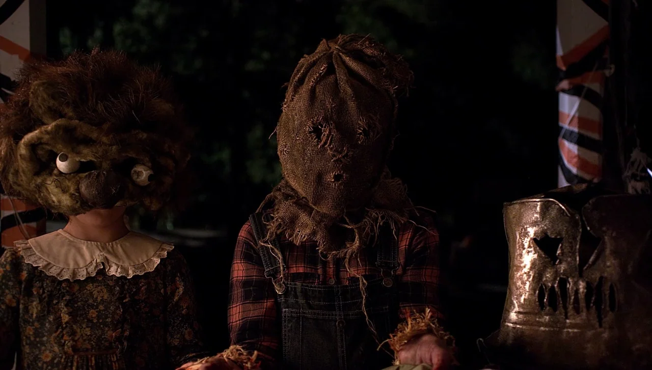 Hellions (2015) movie still: medium — Two children stand on a dark outdoor porch wearing handmade, grotesque halloween masks. The child on the left…