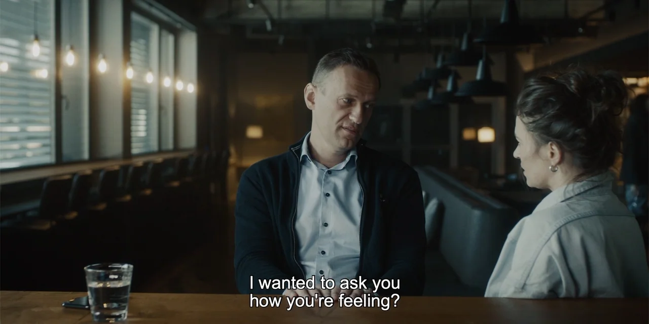 Navalny (2022) movie still: medium — Two individuals sit across from each other at a dimly lit wooden bar counter. A man in a dark sweater talks to…