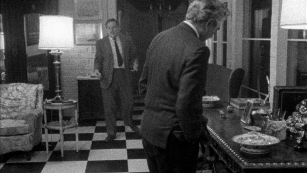 Faces (1968) movie still: medium — A study features checkered tile flooring and a desk cluttered with various decorative objects. Two men in…