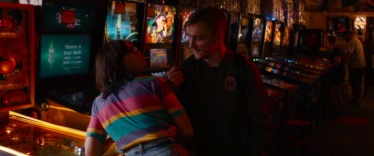 Dinner in America (2020) movie still: medium — A dimly lit arcade interior features rows of glowing pinball machines and silver tinsel decorations.…