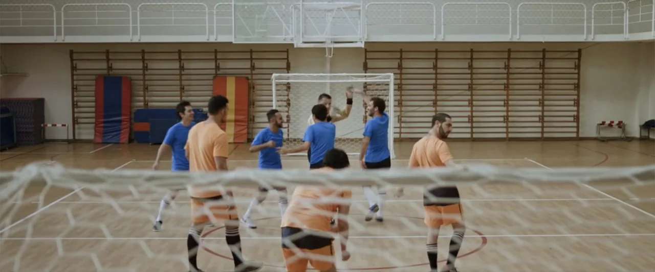 Do It Like An Hombre (2017) movie still: wide — A spacious indoor gymnasium features wooden flooring and wooden wall-mounted bars. Seven men in…