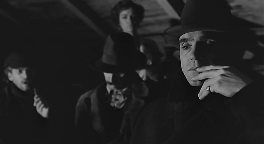 Kafka (1991) movie still: medium — A dimly lit wooden attic space featuring several people huddled in the darkness. A man in the foreground touches…