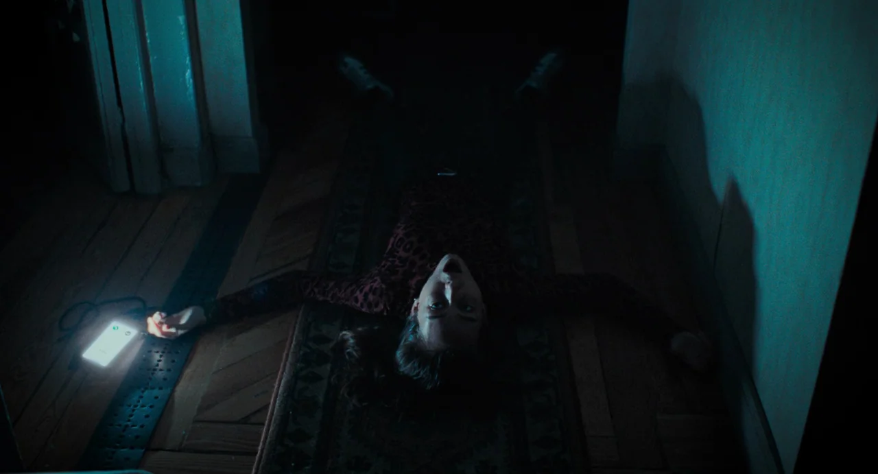 La Abuela (2021) movie still: fullbody — A narrow residential hallway features wooden floors and a dark patterned runner rug. A woman lies flat on…