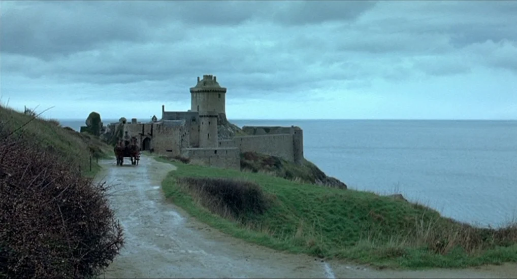 The Last Mistress (2007) movie still: establishing — A stone castle sits on a cliff overlooking the grey sea under overcast skies. A horse-drawn…