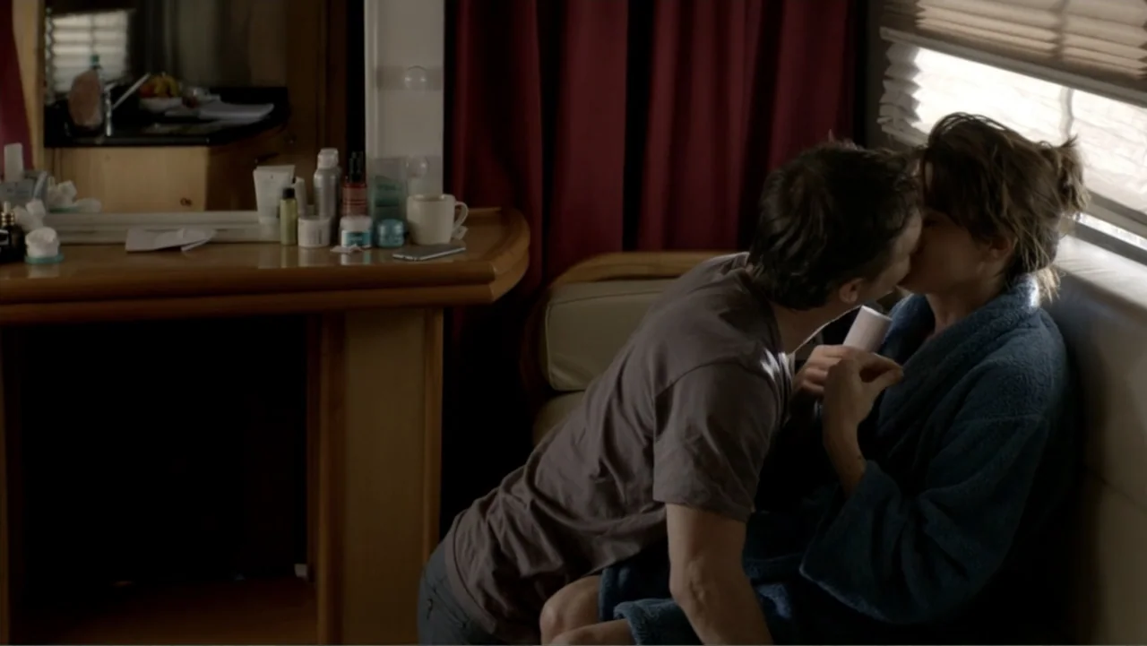 I Married a Dumbass (2016) movie still: medium — A cramped trailer interior features a vanity table with toiletries and dark red curtains. A man in…