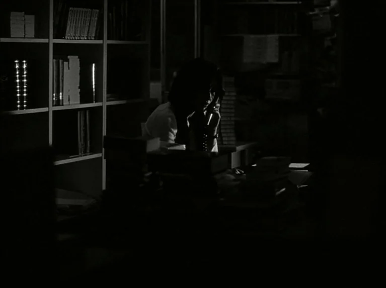 Eloge de L’amour (2001) movie still: medium — A dark, quiet space filled with floor-to-ceiling shelves packed with books. A woman sits at a…