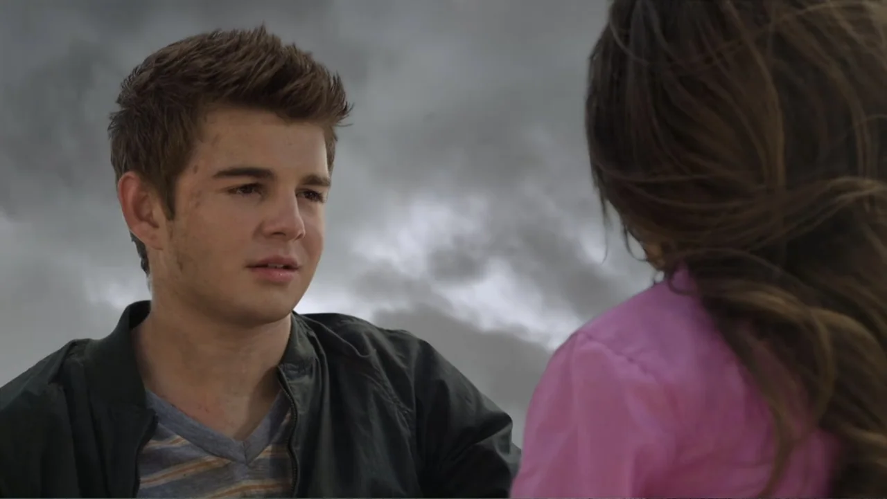 Jinxed (2013) movie still: medium — An open outdoor space is covered by a dense, overcast grey sky. A young man sits opposite a girl whose back is…