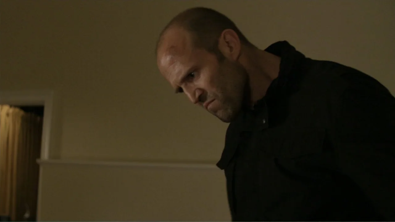 Parker (2013) movie still: medium — A dimly lit indoor space features plain walls and a dark doorway in the background. A man stands in the…