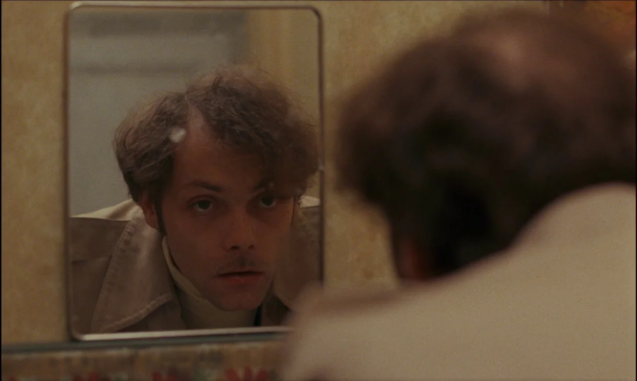 Serie Noire (1979) movie still: closeup — A dimly lit room with yellow patterned wallpaper visible behind a small rectangular mirror. A man gazes…