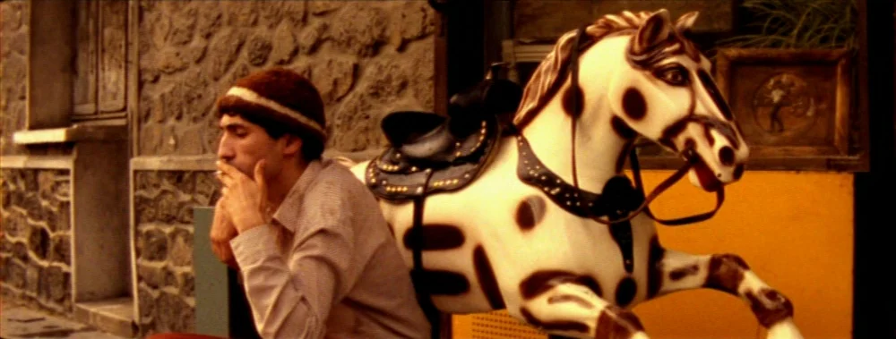 Carne (1991) movie still: medium — A stone building with rough-hewn masonry serves as a backdrop on a city sidewalk. A man in a knit cap sits beside…