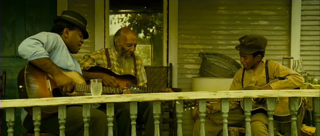 I’m Not There (2007) movie still: medium — Three men play guitars on the wooden porch of a house with light siding. A metal basin rests in the…