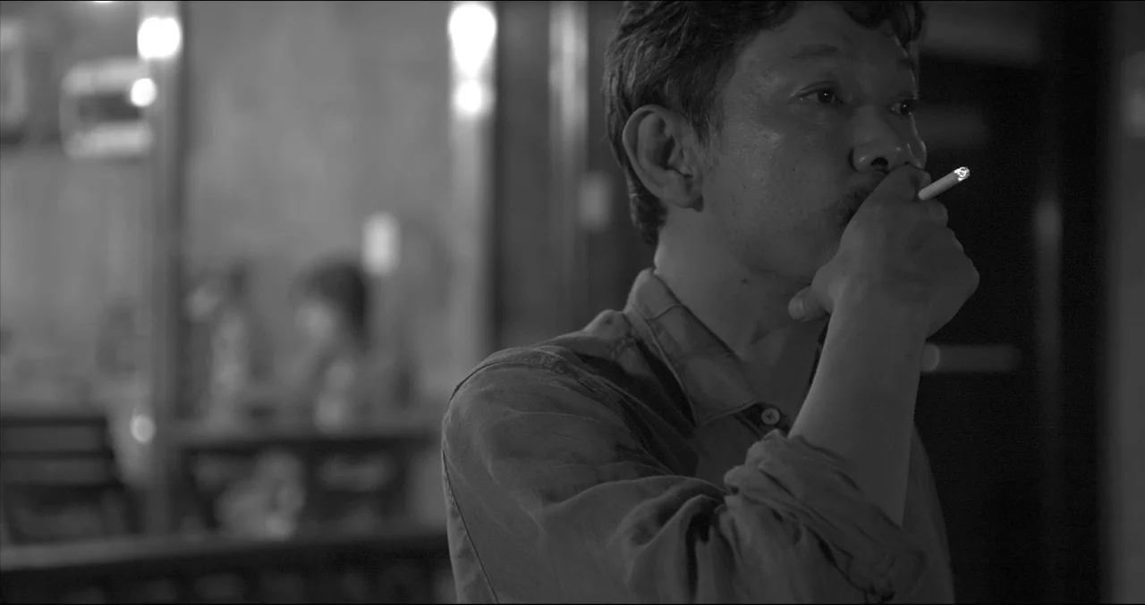 Grass (2018) movie still: medium — A dark indoor space with blurred furniture in the background. A man holds a lit cigarette while looking off-camera.