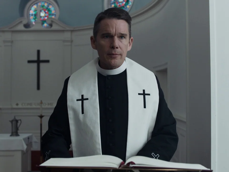 First Reformed (2017) movie still: medium — A stark, minimalist white sanctuary features simple wooden crosses mounted on the walls. A priest in…