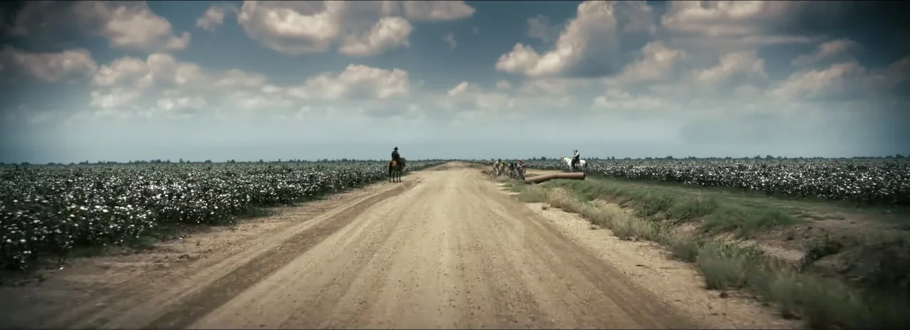 Sinners (2025) movie still: wide — A long, flat dirt road bisects large fields of white cotton under a cloudy sky. Horse riders and people move away…