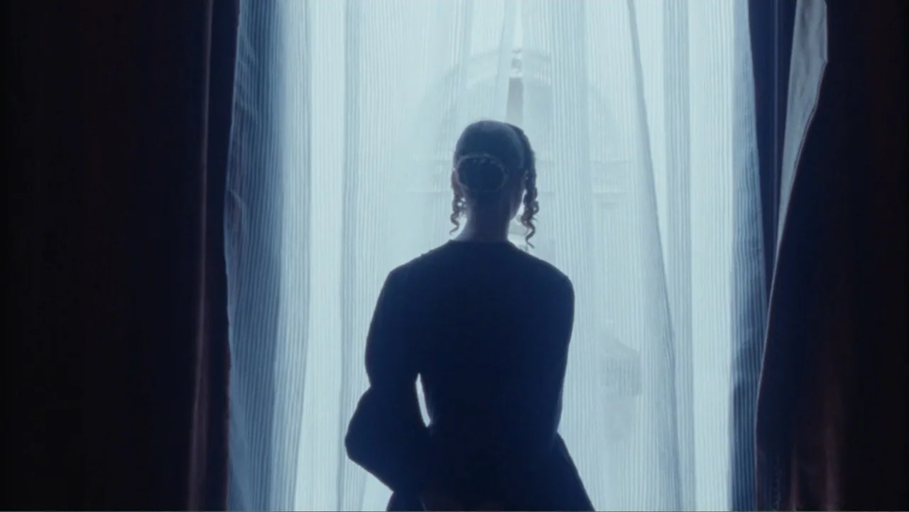 Demon in the White House (2021) movie still: medium — An interior room with heavy curtains framing a bright window overlooking a building exterior.…