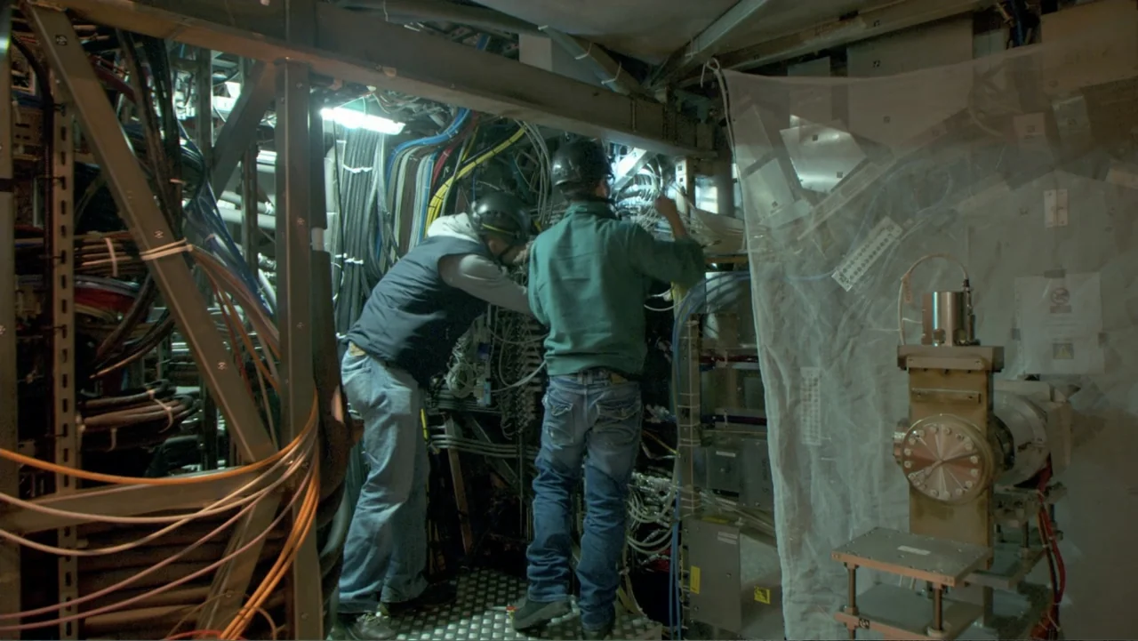 CERN (2013) movie still: medium — A technical facility interior filled with bundles of cables and metallic equipment racks. Two workers in hard hats…