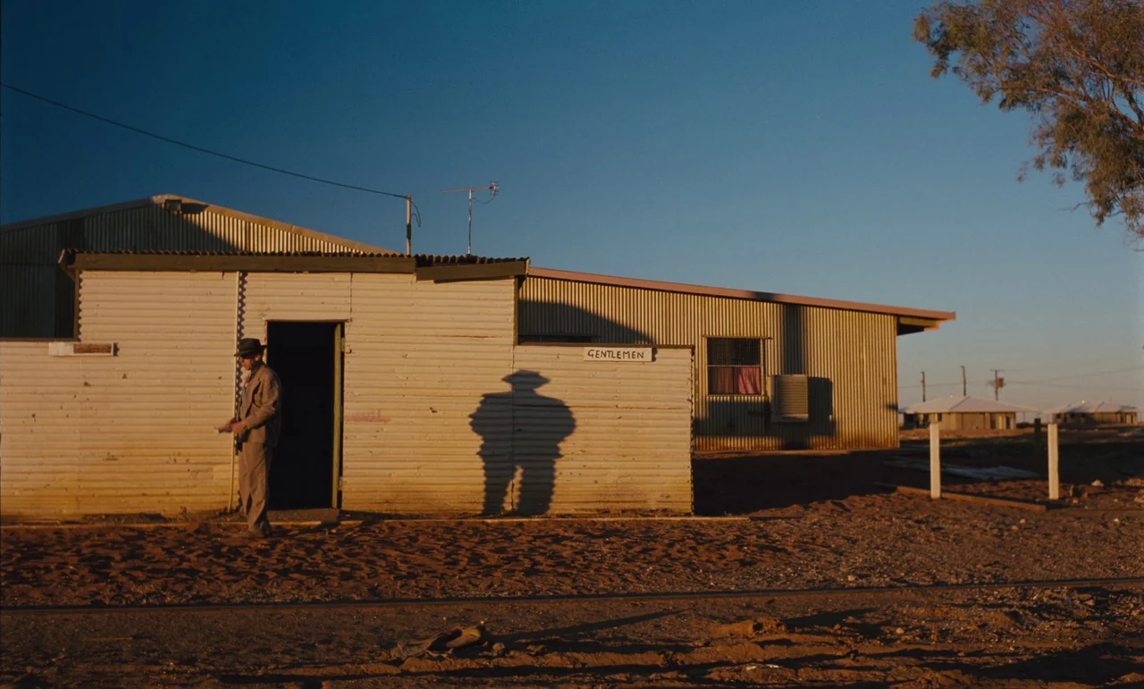 Until The End Of The World (1991) movie still: wide — A corrugated metal building with a 'GENTLEMEN' sign sits on dry, reddish dirt ground. A man…