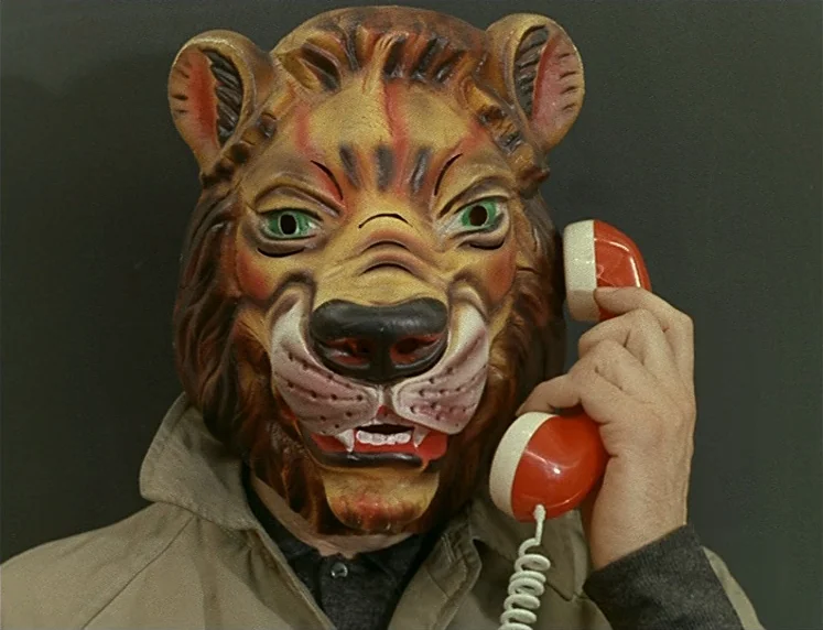 La Chinoise (1967) movie still: closeup — A person wears a vintage molded plastic lion mask against a dark wall background. They hold a red vintage…