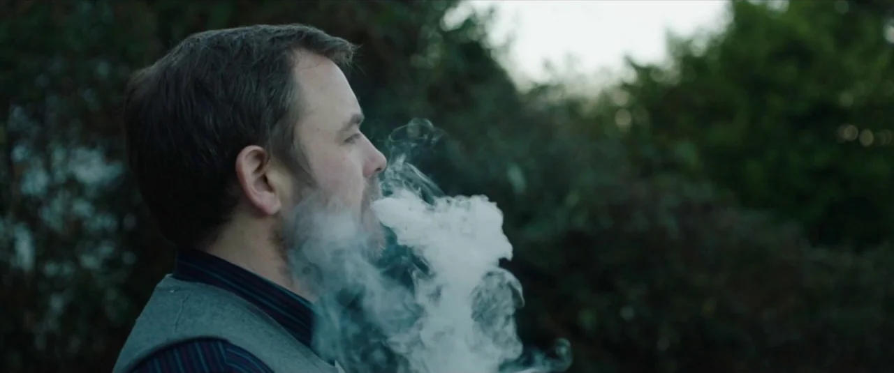 Happy New Year, Colin Burstead (2018) movie still: closeup — A man in a sweater vest stands outdoors among dense green foliage. He exhales a dense…