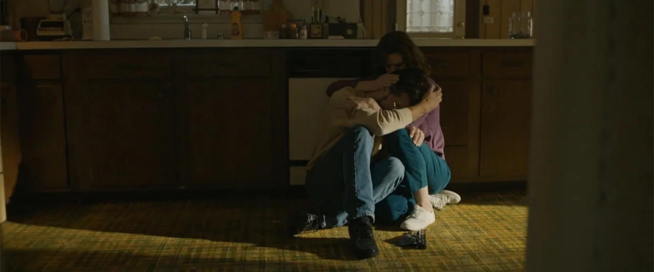 Unsung Hero (2024) movie still: fullbody — A residential room features dark wood cabinetry and patterned carpet flooring. A man and a woman sit…