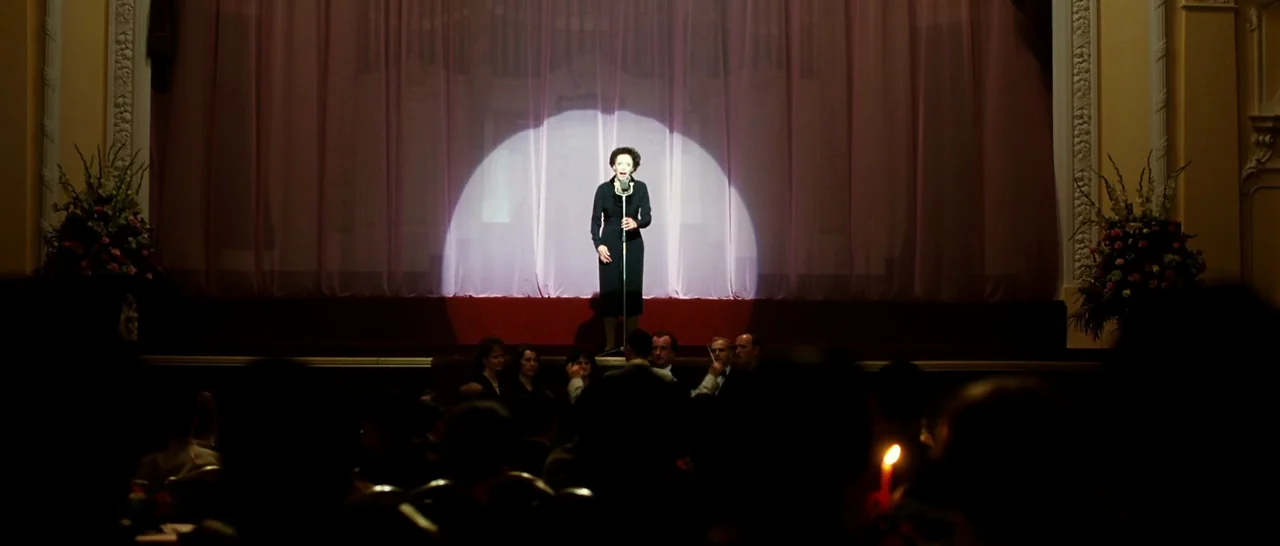 La Vie En Rose (2007) movie still: wide — An illuminated singer stands center on a wooden stage in front of pleated pink curtains. The darkened…