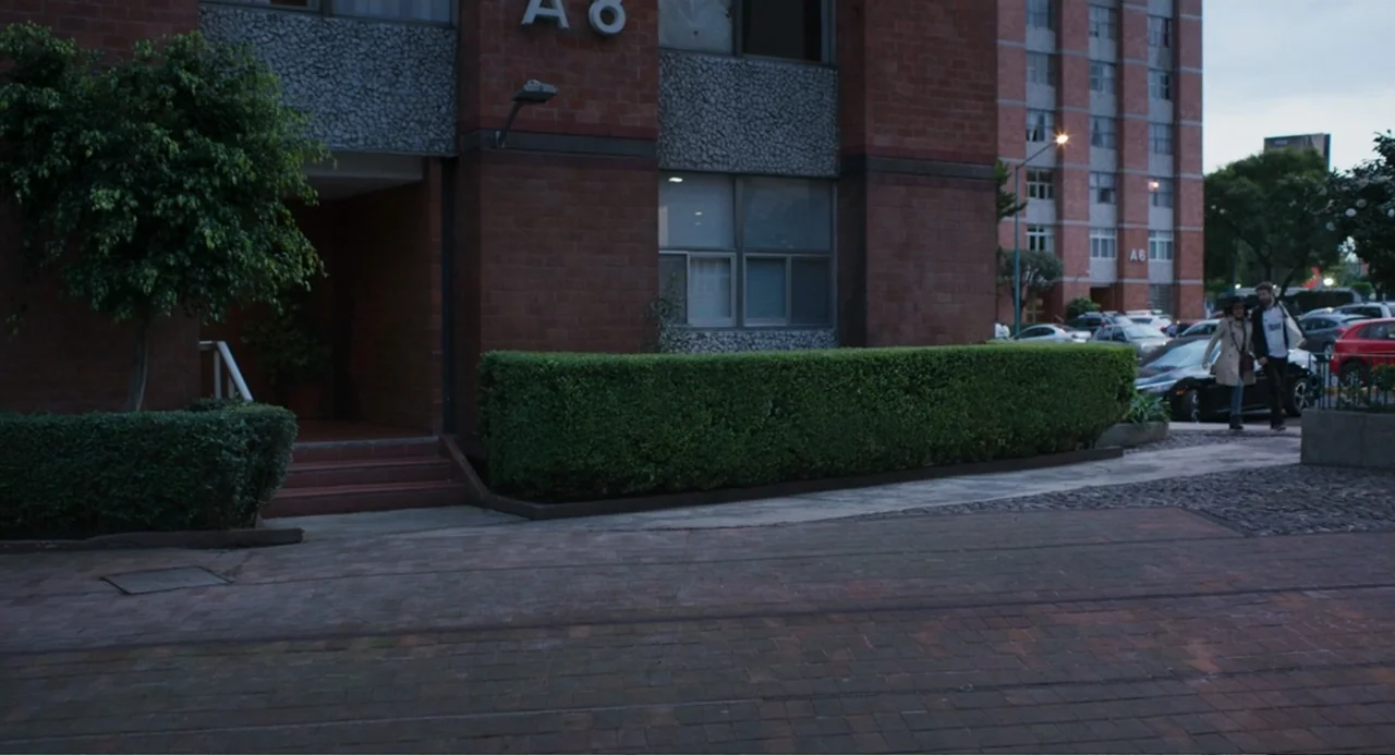 The Lift (2021) movie still: wide — A multi-story brick apartment building features a manicured hedge and a paved walkway. A young man and a woman…