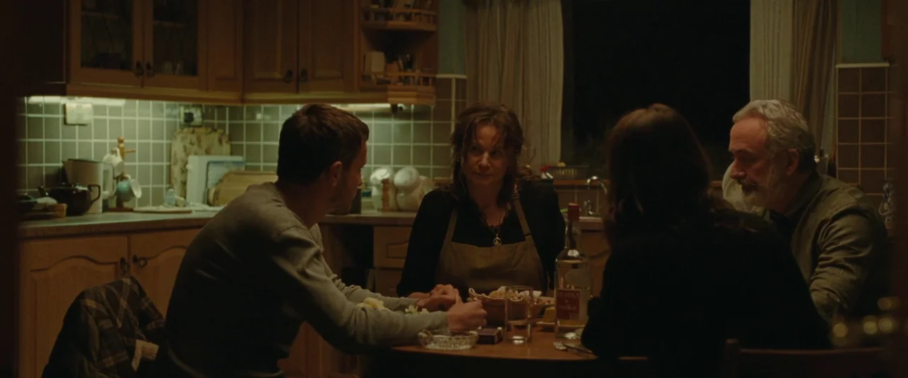 God’s Creatures (2022) movie still: medium — A residential kitchen features wooden cabinetry and light-colored square tile walls. Four adults sit…