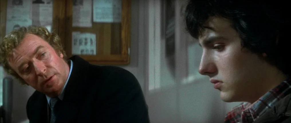 Dressed to Kill (1980) movie still: medium — A room with paper notices pinned to a wooden bulletin board on the wall. An older man in a suit speaks…
