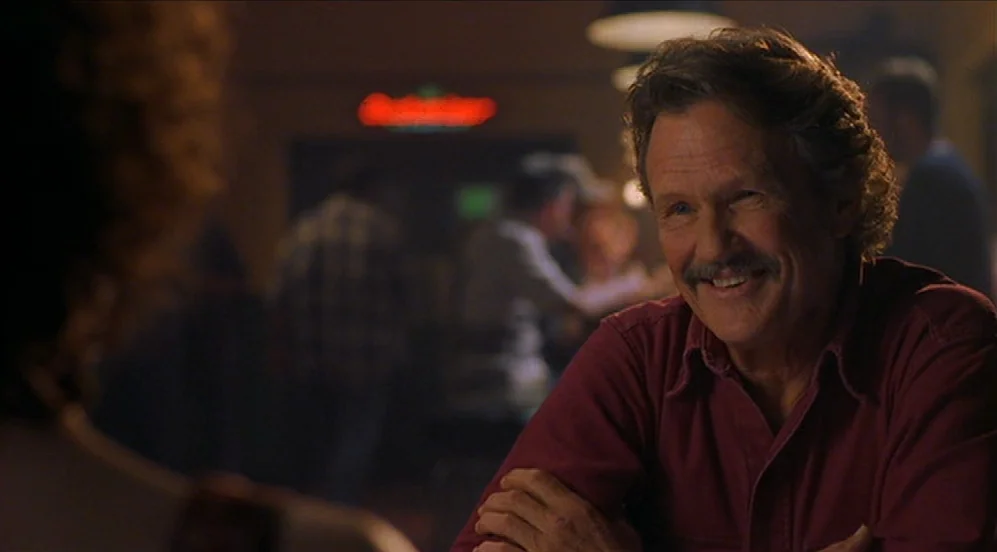 Limbo (1999) movie still: medium — A dimly lit bar with patrons visible in the soft-focus background. A middle-aged man smiles while sitting with…
