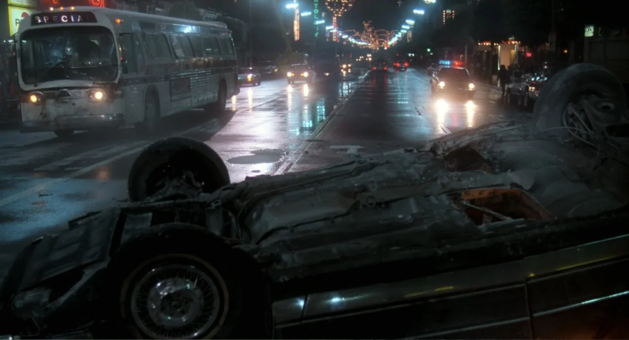 Lethal Weapon (1987) movie still: wide — An overturned sedan lies on its roof in the center of a wet city thoroughfare. In the background a city bus…
