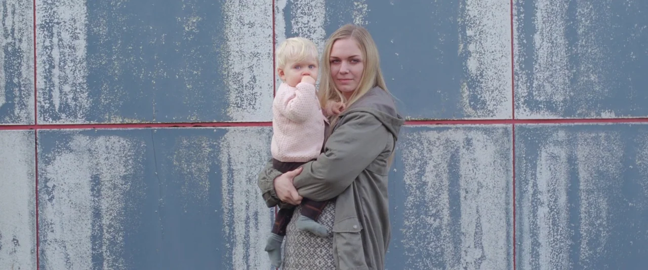 A White, White Day (2019) movie still: medium — A woman stands against a textured gray exterior wall with red vertical joints. She holds a young…