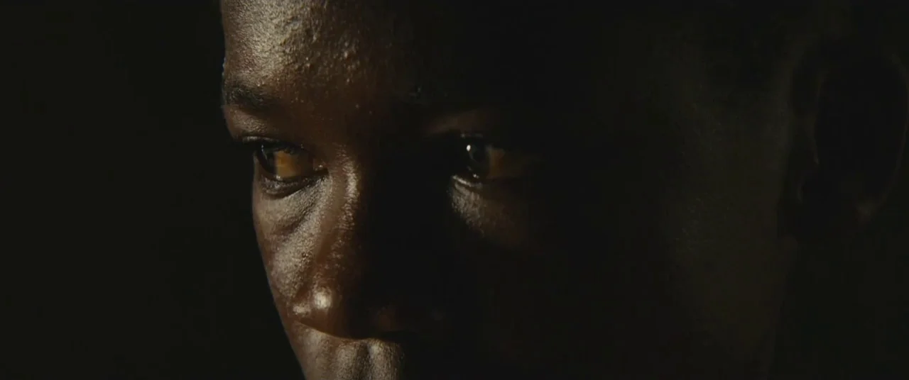 Beasts of No Nation (2015) movie still: closeup — A dark, underexposed environment leaves only the face visible. A young Black man stares intently…