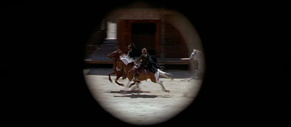 For A Few Dollars More (1965) movie still: wide — A dusty outdoor yard is framed through a circular telescope or binoculars lens. Two men ride…