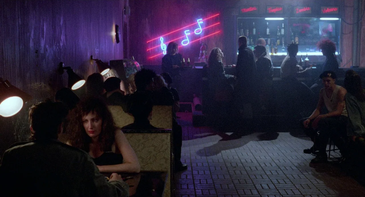 Sid and Nancy (1986) movie still: wide — A dimly lit 1980s lounge features purple walls and a prominent pink neon musical sign. Patrons occupy…