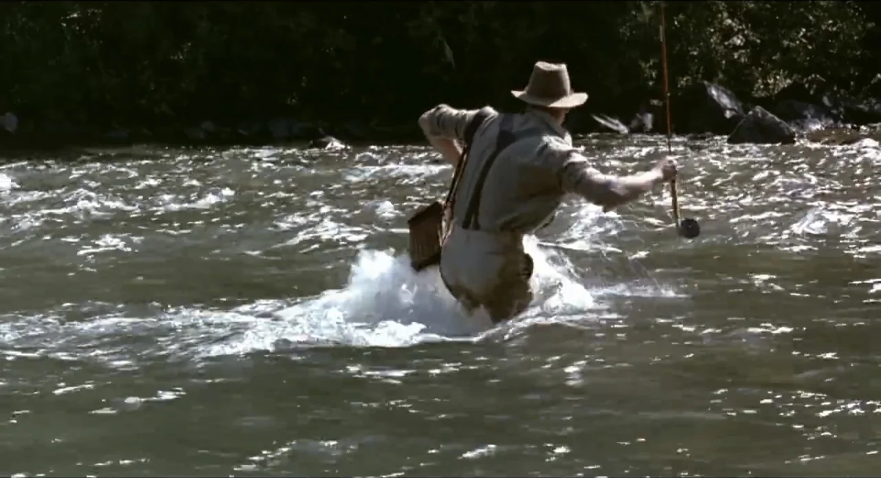 A River Runs Through It (1992) movie still: medium — A shallow river with rapids flowing over dark rocks and vegetation on the bank. A man in…