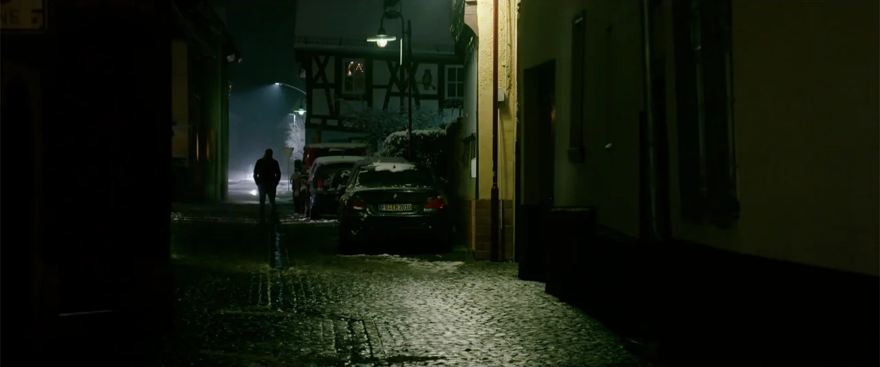 Simple as Water (2021) movie still: wide — A narrow cobblestone street at night is covered with patches of melting winter snow. A lone person in a…