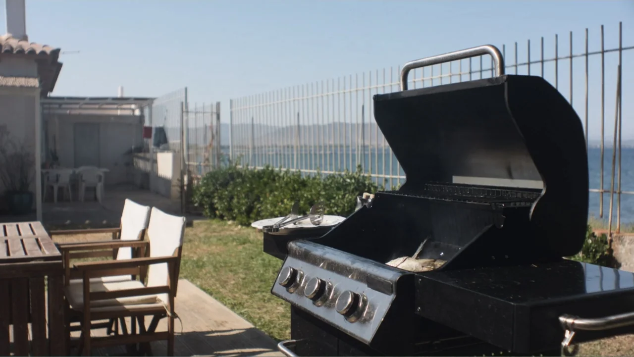 Hot Milk (2025) movie still: medium — A residential patio overlooks a coastal bay with a metal fence separating the yard. An open gas grill stands…