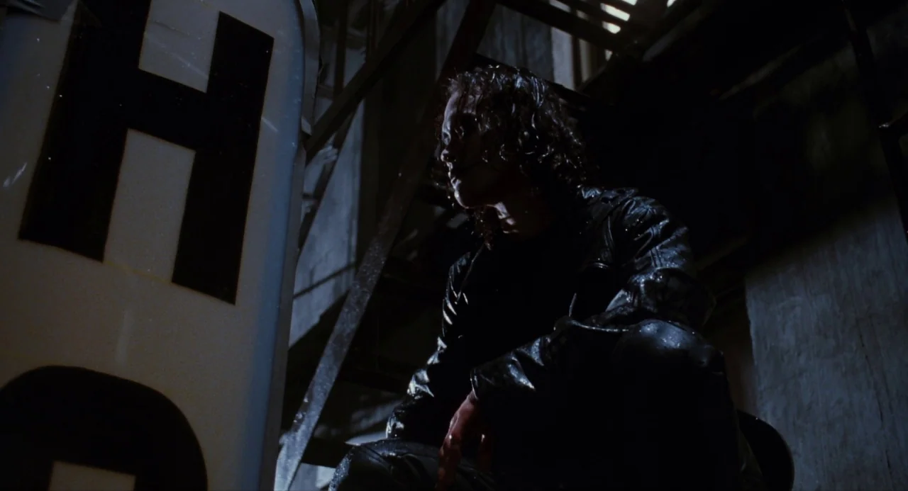 The Crow (1994) movie still: medium — A dark fire escape at night features an illuminated neon sign fragment. A man in a wet leather jacket crouches…