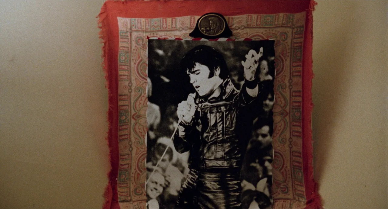 Out of the Blue (1980) movie still: closeup — A square print of Elvis Presley is displayed on a decorative, paisley-patterned textile background.…