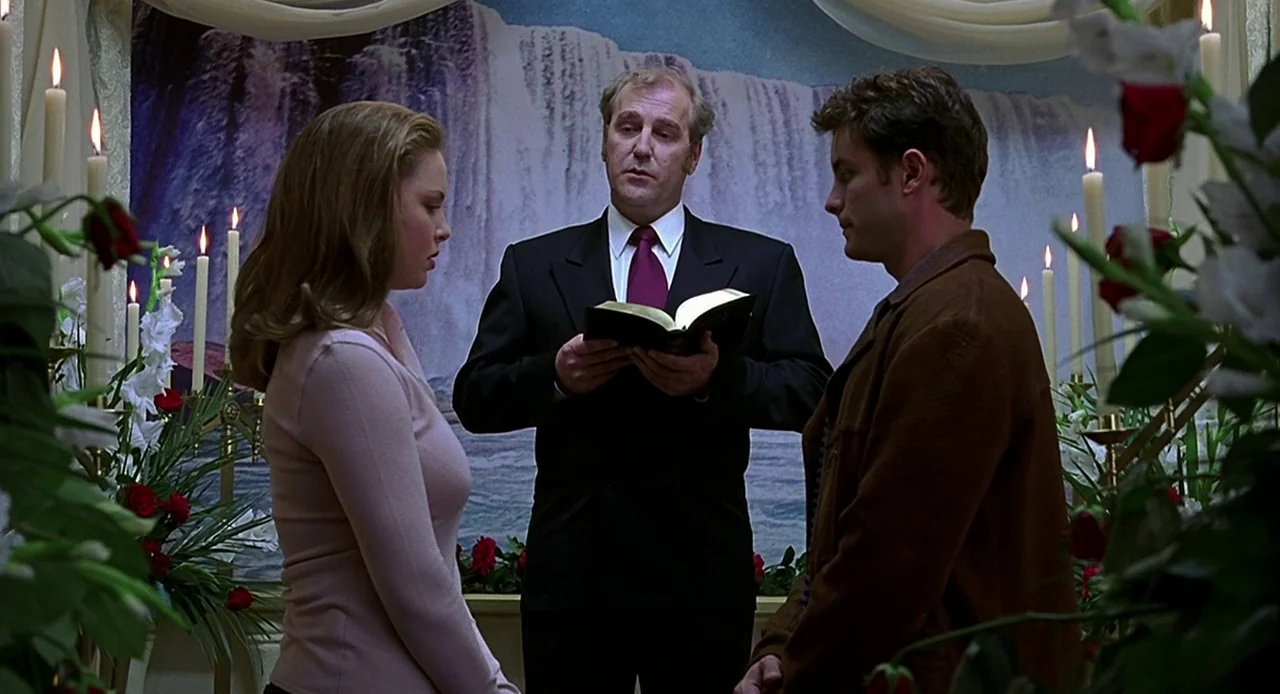 Bride of Chucky (1998) movie still: medium — A small wedding ceremony occurs in front of a waterfall mural with floral arrangements. An officiant…