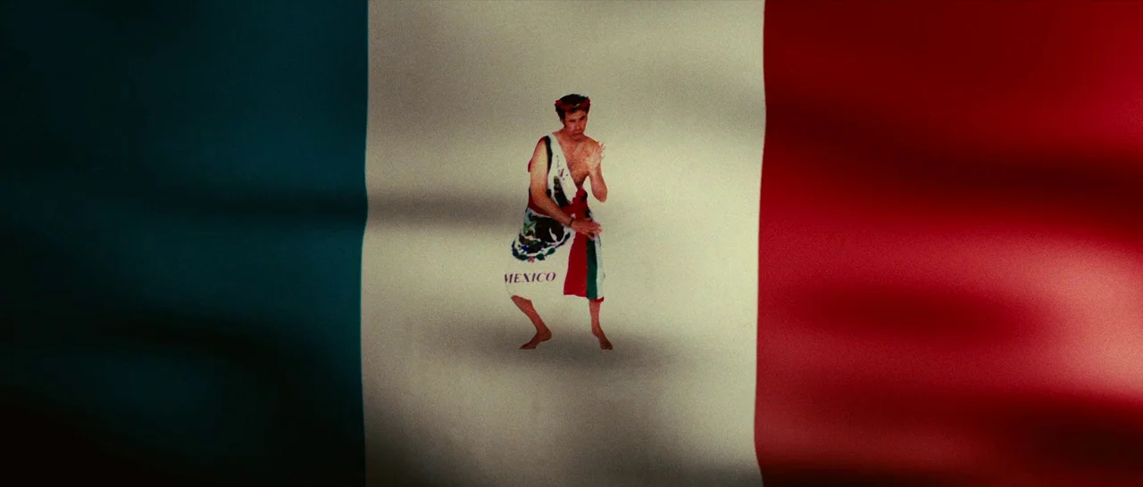 Casa De Mi Padre (2012) movie still: wide — A man stands centered against the large, waving fabric of the Mexican national flag. The background is…