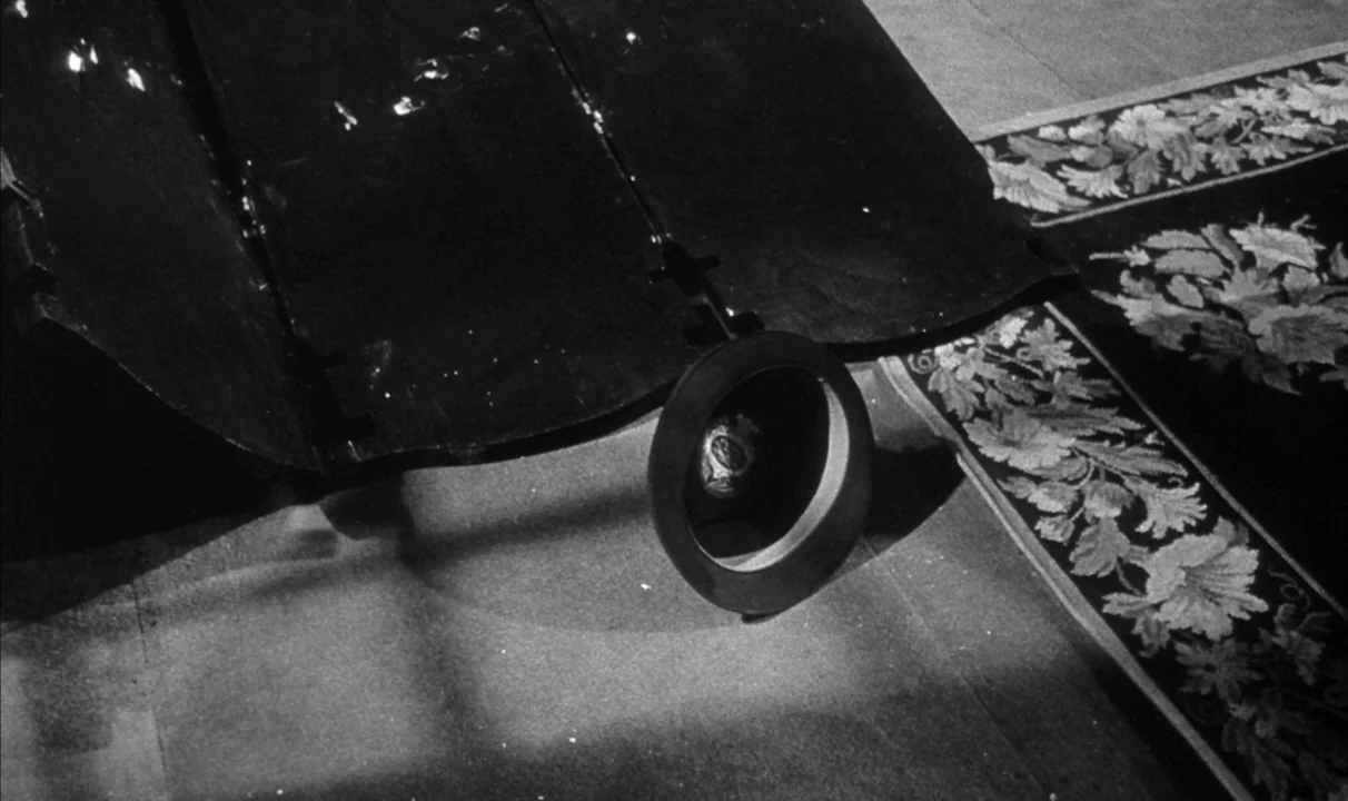 Le Doulos (1962) movie still: closeup — A dark, upside-down top hat sits on a floor covered by an ornate floral rug. The hat is illuminated by a…