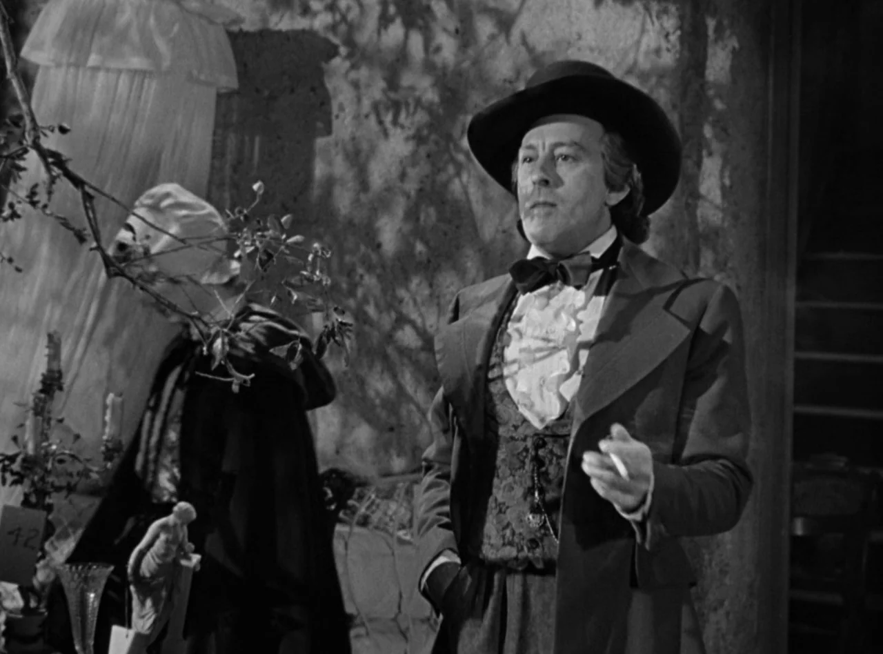 Madame Bovary (1949) movie still: medium — An indoor room contains a cluttered backdrop with furniture and decorative objects. A middle-aged man…