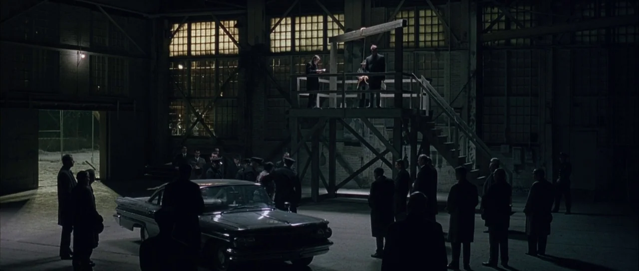 Capote (2005) movie still: wide — A dark, cavernous industrial space features a wooden gallows platform constructed on the floor. A gathering of men…