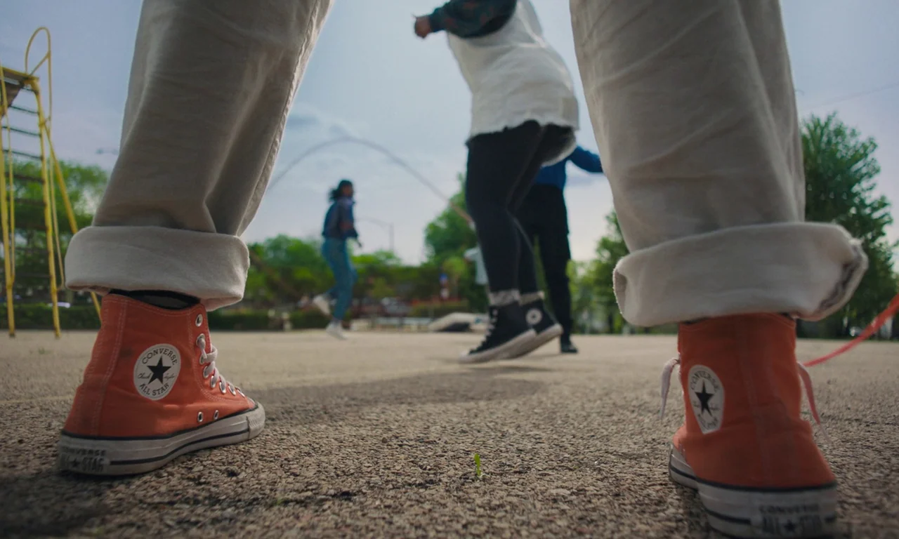 We Grown Now (2023) movie still: wide — An asphalt playground ground-level view focuses on a person's bright orange sneakers. People in the…