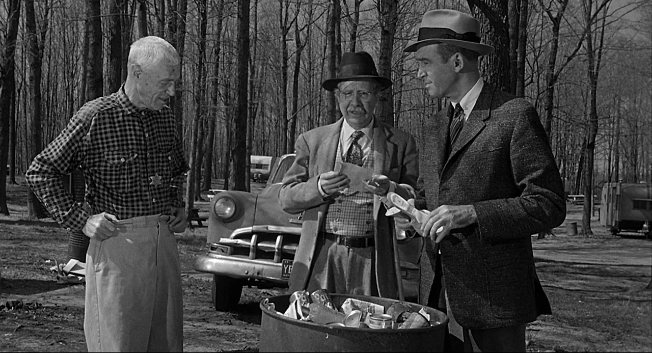Anatomy of a Murder (1959) movie still: medium — A wooded campsite contains a classic 1950s automobile and a large metal trash barrel. Three men…