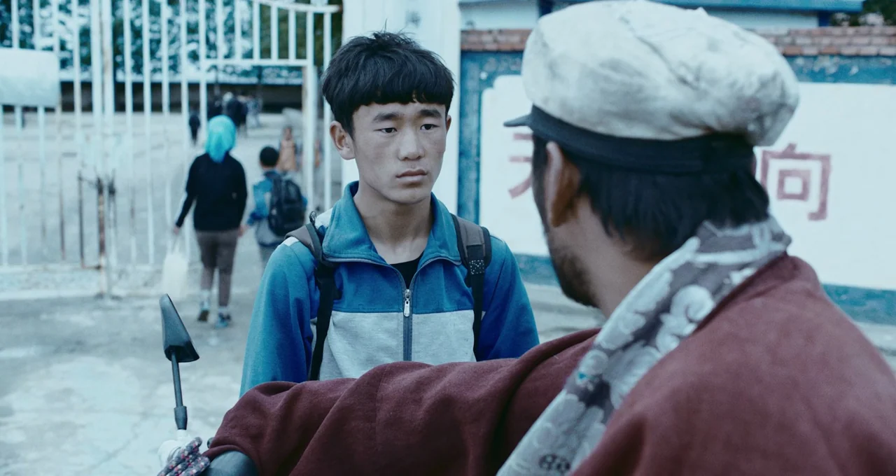 Balloon (2019) movie still: medium — An outdoor area features a white metal gate and a painted wall with characters. A young man stands facing an…