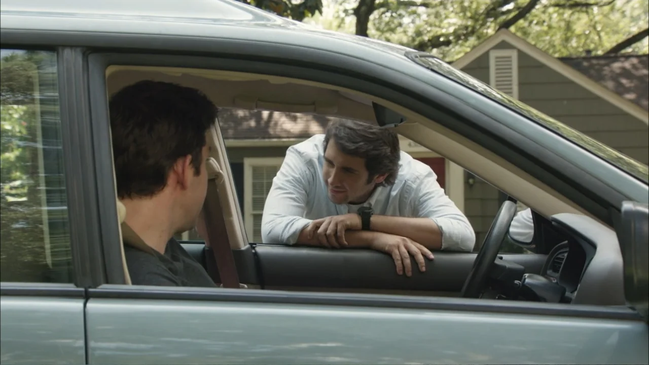 The Hollars (2016) movie still: medium — A sunny suburban driveway with a house visible in the soft-focus background. One man leans on the door of a…
