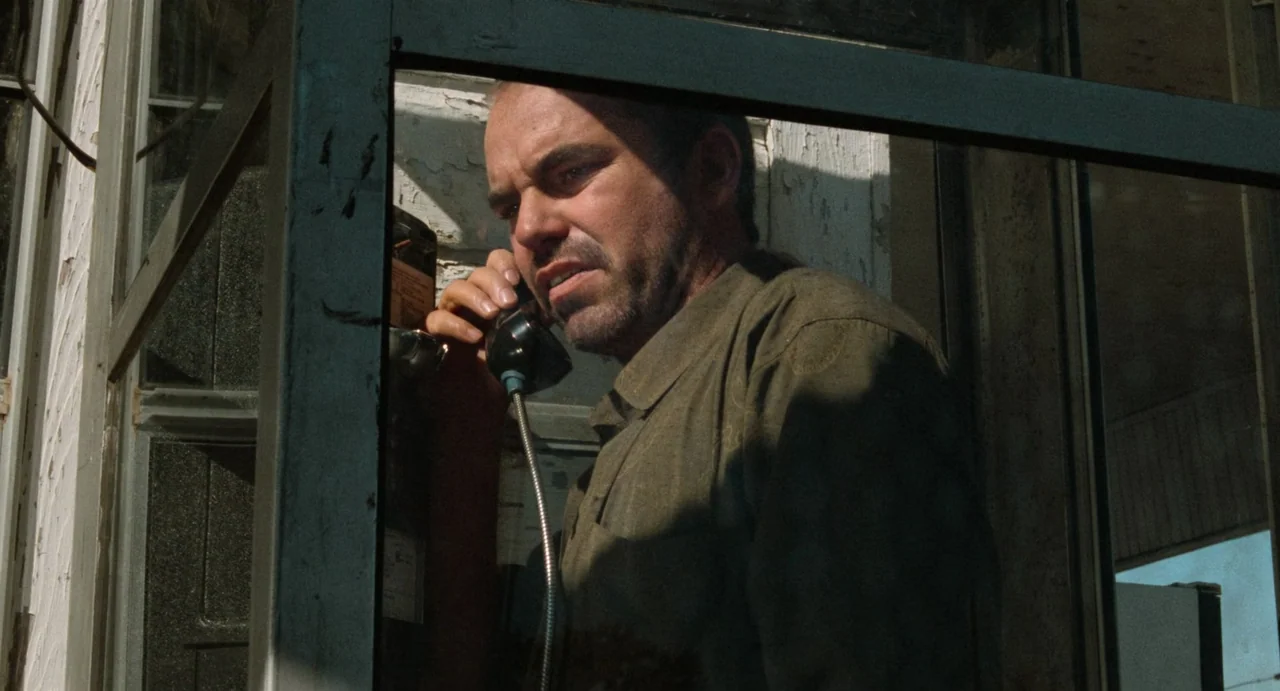 One False Move (1991) movie still: medium — A man stands inside a glass-paneled outdoor telephone enclosure during the day. He holds a black…